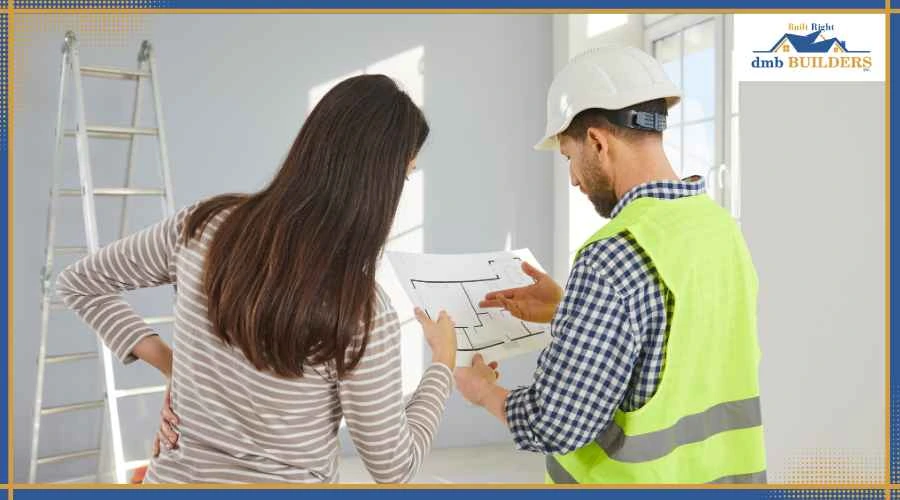 Woman and construction worker in hard hats review a blueprint indoors with a ladder in the background and safety gear visible.